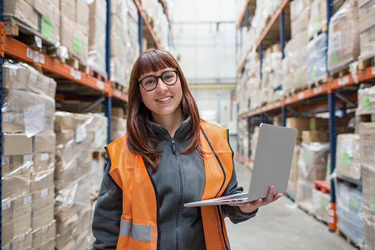Woman in a warehouse, wearing safety vest and holding laptop, keeping track of inventory and business sales and use tax